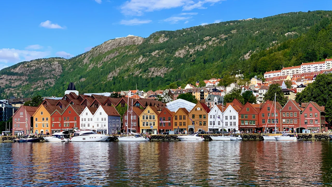 Värikkäät historialliset rakennukset Bryggenissä Bergenissä veden äärellä veneiden kanssa, vihreä vuori sinisen taivaan alla.