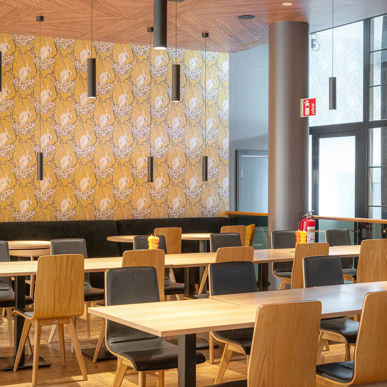 Wooden tables and chairs sit in a café with vibrant yellow floral wallpaper. Black pendant lights hang from the ceiling. A fire extinguisher and emergency sign are visible.