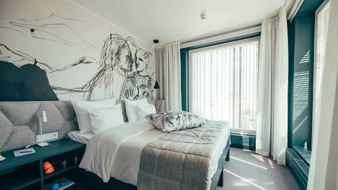 En pent sengered med hvitt sengetøy og en mønstret pute står i et rom med et stort veggmaleri, ved siden av et vindu med tynne gardiner.