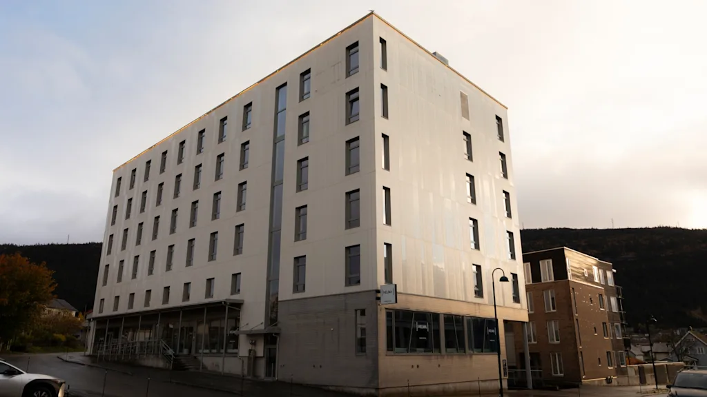 En moderne fleretagers bygning med mange vinduer og skilt Helma, står ved en gade med biler. Bakker i baggrunden.