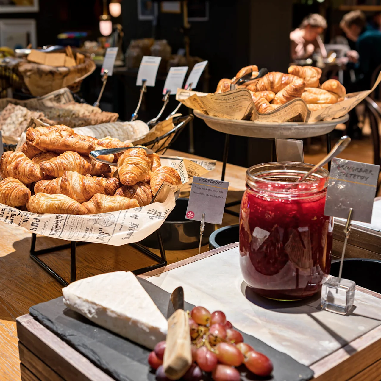 Croissanter vises på en buffé med jordbær- og appelsinmarmelade, druer og assorterede brød. Spisende gæster ses i baggrunden af restaurantmiljøet.