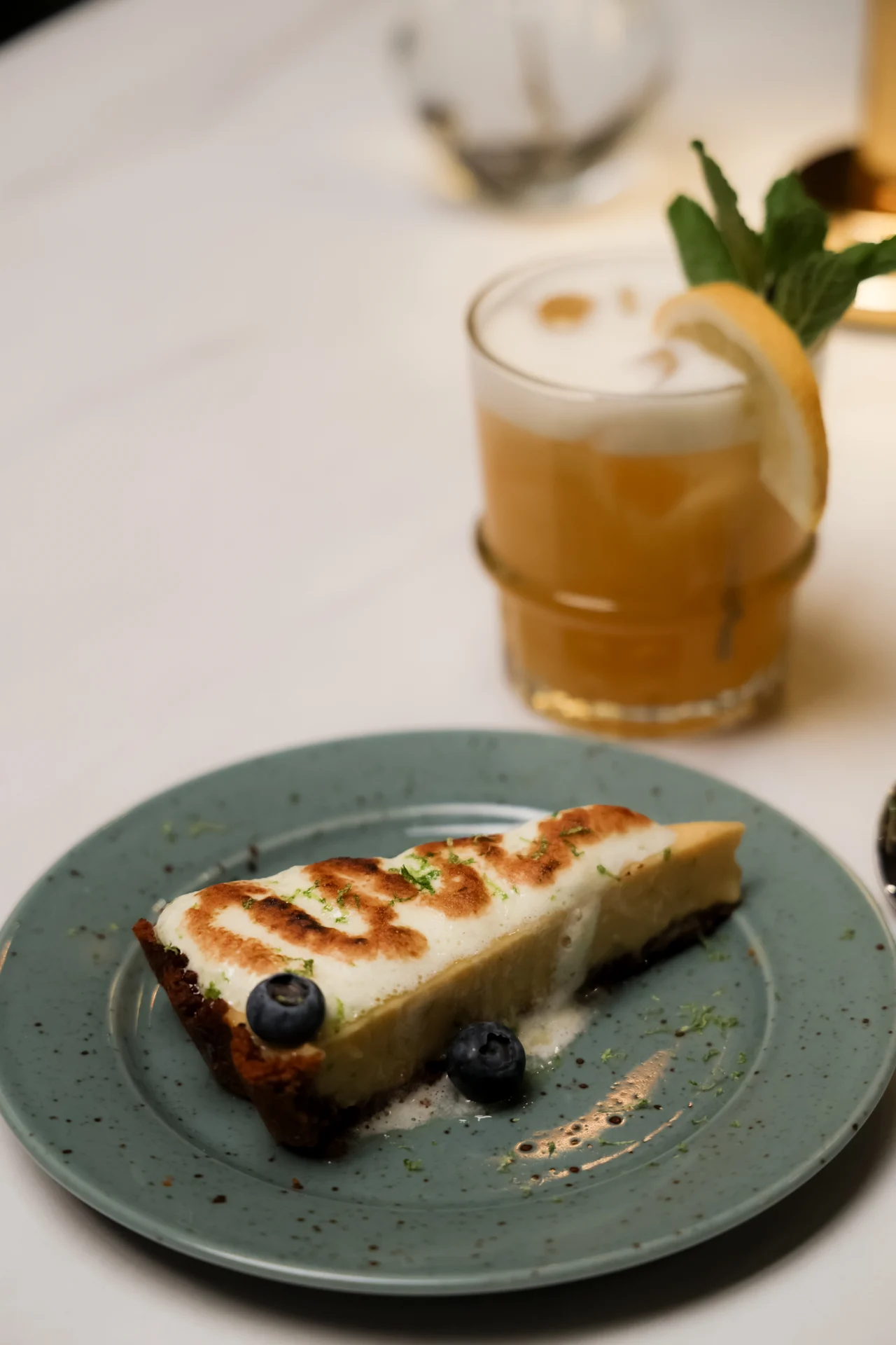 A slice of pie with a toasted top and blueberries sits on a green plate. Nearby, a frothy orange drink garnished with lemon and mint is on a table.