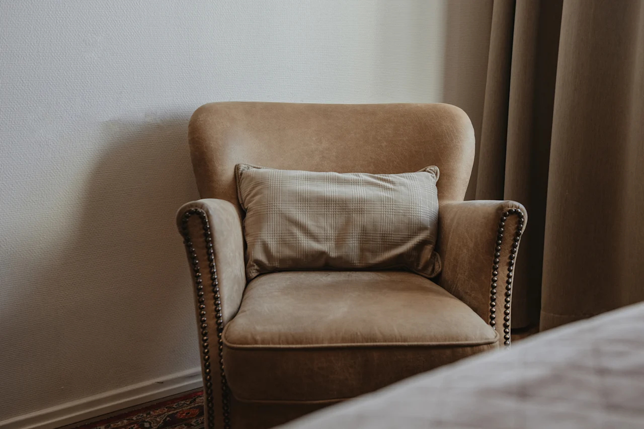 En mysig, ljusbrun fåtölj med en rutig kudde står i ett rum och inbjuder till avkoppling bredvid ett fönster med gardiner.