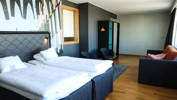Två prydligt bäddade enkelsängar står sida vid sida mot en dekorativ vägg i ett rymligt hotellrum. I närheten kompletterar moderna stolar och en soffa trägolvet.