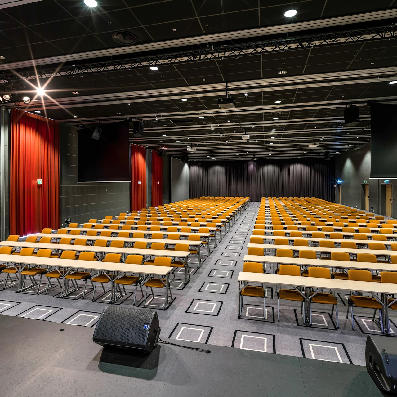 Rows of empty yellow chairs and long tables are aligned in a large auditorium with red curtains and dark ceiling, creating a formal setting.