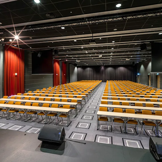 Rader med tomme gule stoler og lange bord er oppstilt i et stort auditorium med røde gardiner og mørkt tak, som skaper en formell atmosfære.
