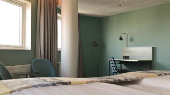 Et skrivebord med lamper står mod en grøn væg, en mønstret stol foran. I nærheden er et vindue med grå gardiner og en søjle, der giver dybde til rummet.