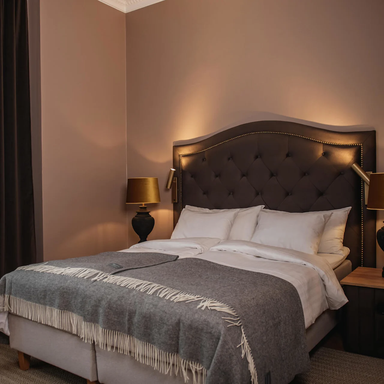 A neatly made bed with a tufted headboard, flanked by two dark lamps, rests in a warmly lit bedroom with brown walls and heavy curtains.