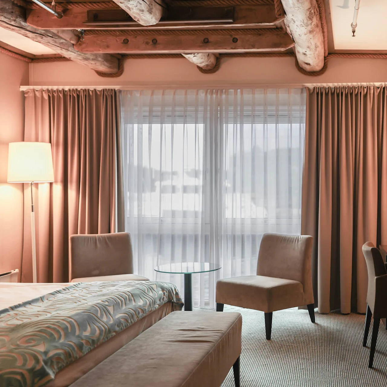 A hotel room with a bed and decorative blanket sits under wooden beams. Adjacent, a desk with a lamp and television faces a window draped with curtains. 