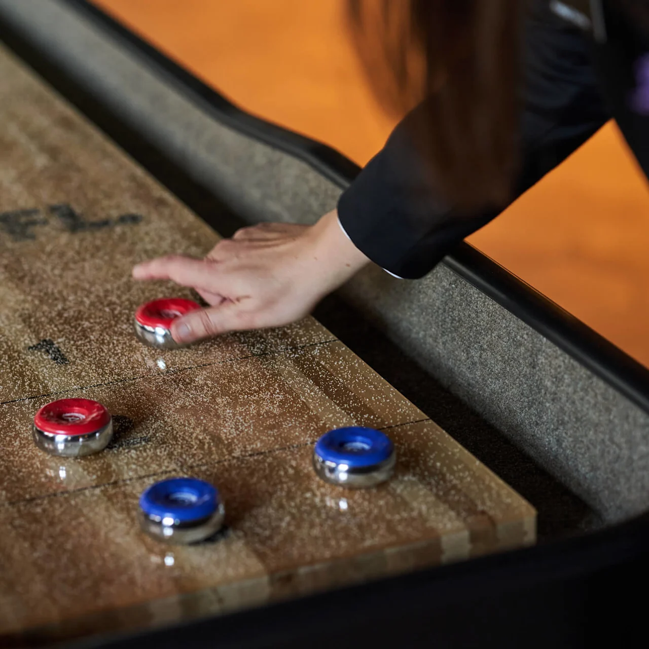 En hånd skubber en rød puck på et shuffleboard-bord af træ, med blå og røde pucke spredt; teksten "SHUFL." på overfladen.