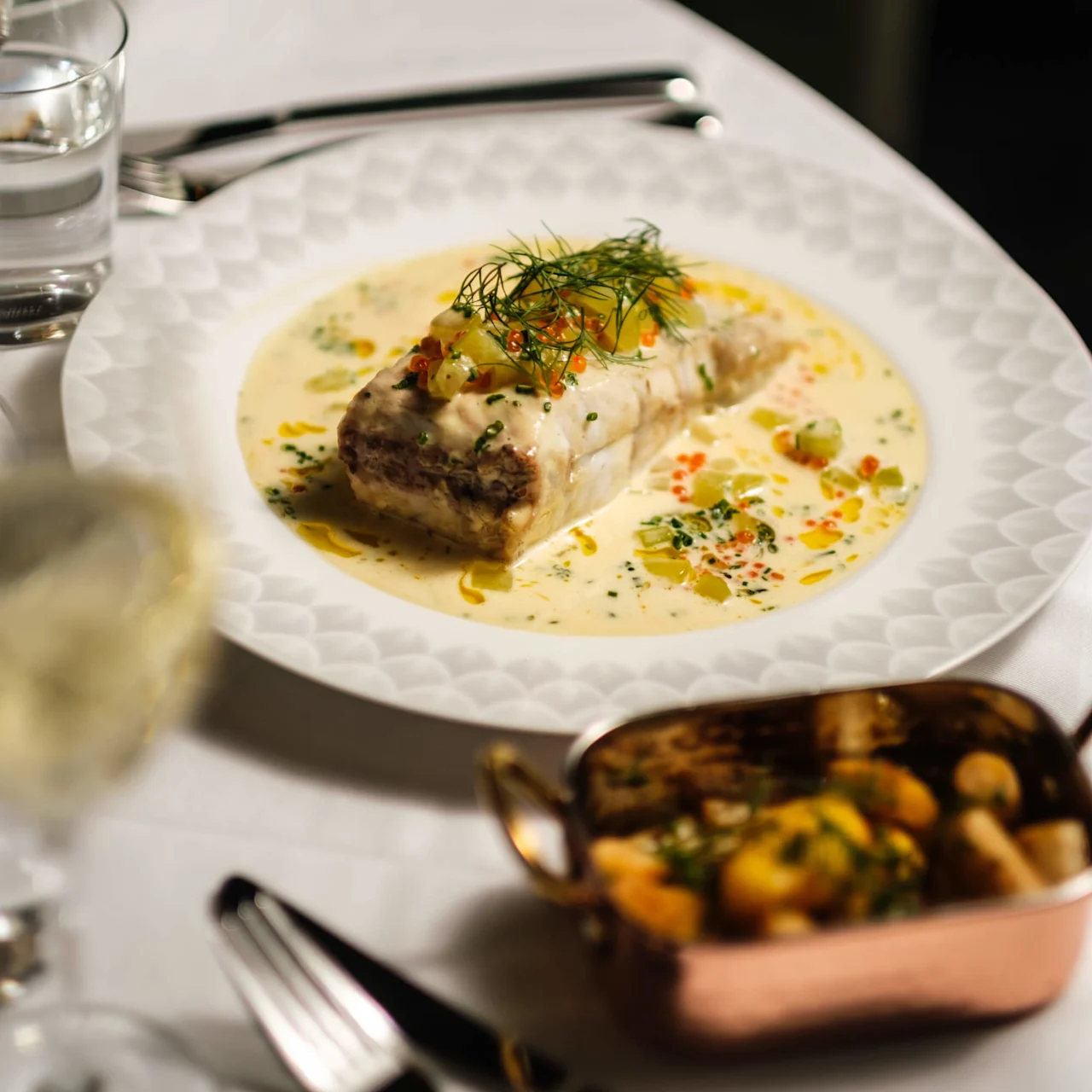 En tallrik med gourmeträtt av fisk i krämig sås toppad med örter, serveras på en vit duk bredvid en skål med rostade grönsaker och ett glas vatten.