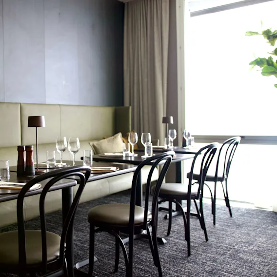 Ett restaurangbord dukat med vinglas och bestick, placerat på ett mörkt golv nära en ljusgrön vadderad sittplats, under mjuk belysning i ett modernt matområde.