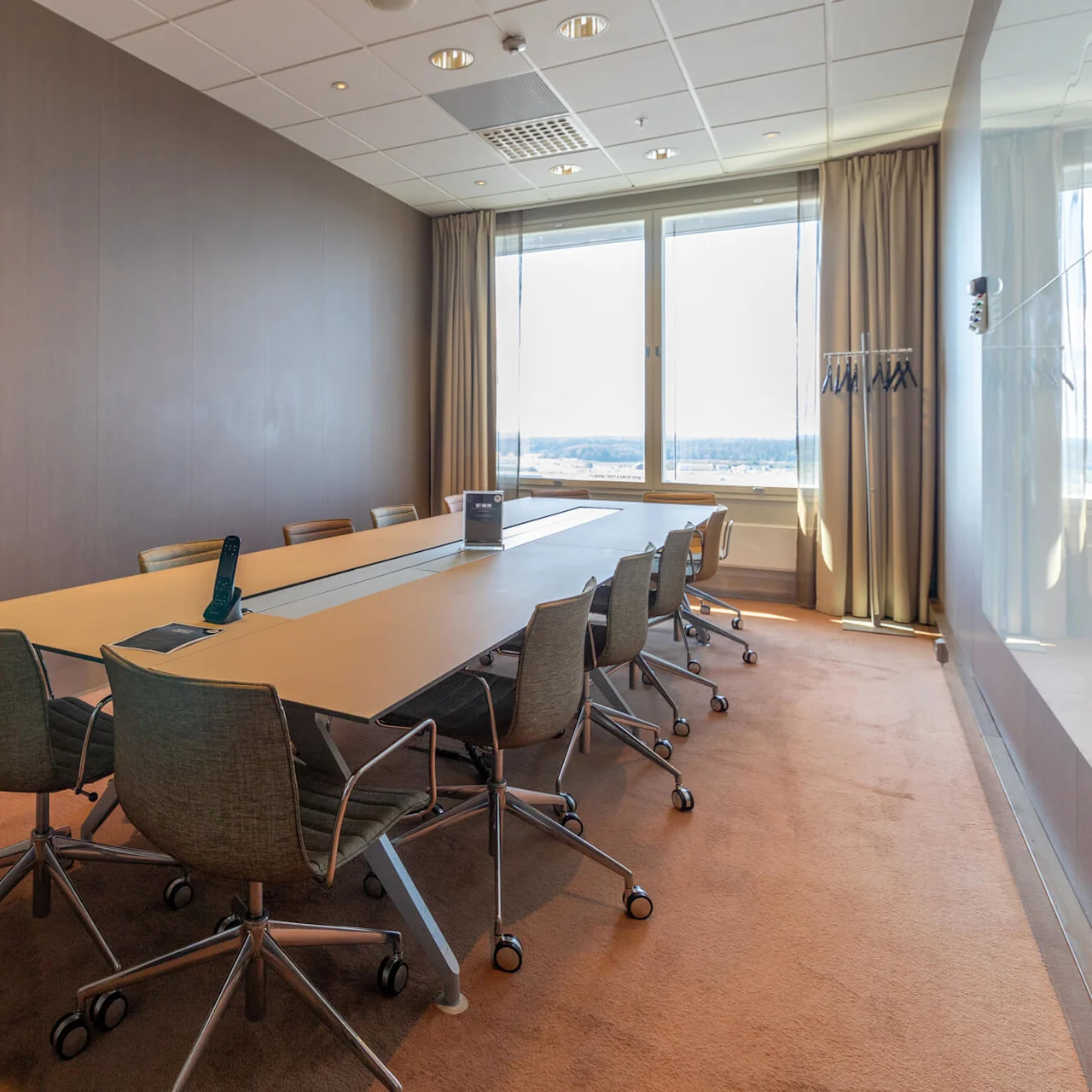 A long conference table with chairs surrounds it; a remote sits on the table. The room features large windows with a view, beige carpet, and minimal decor.