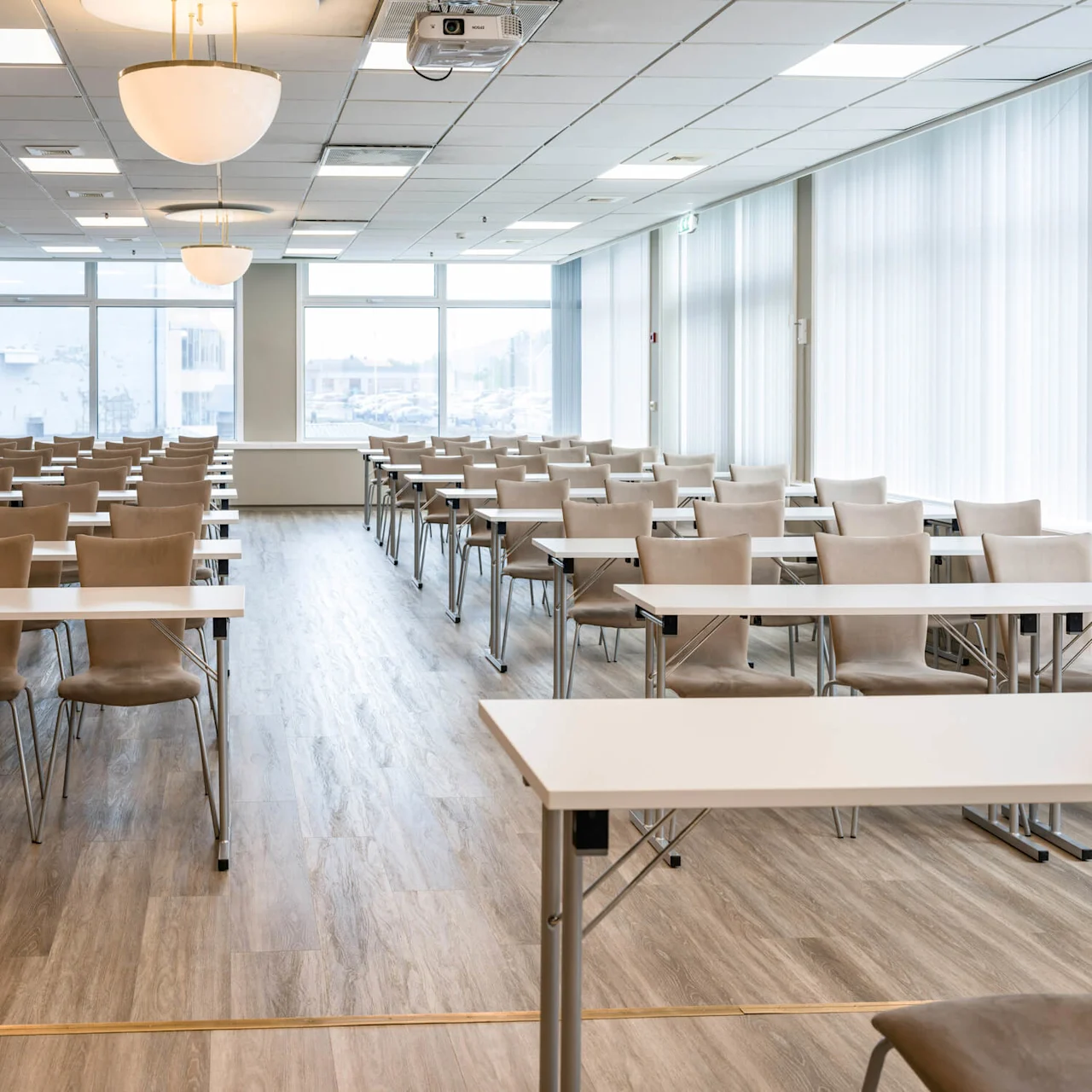 Empty beige chairs are arranged in neat rows behind white tables in a spacious, well-lit conference room with large windows and wooden flooring.