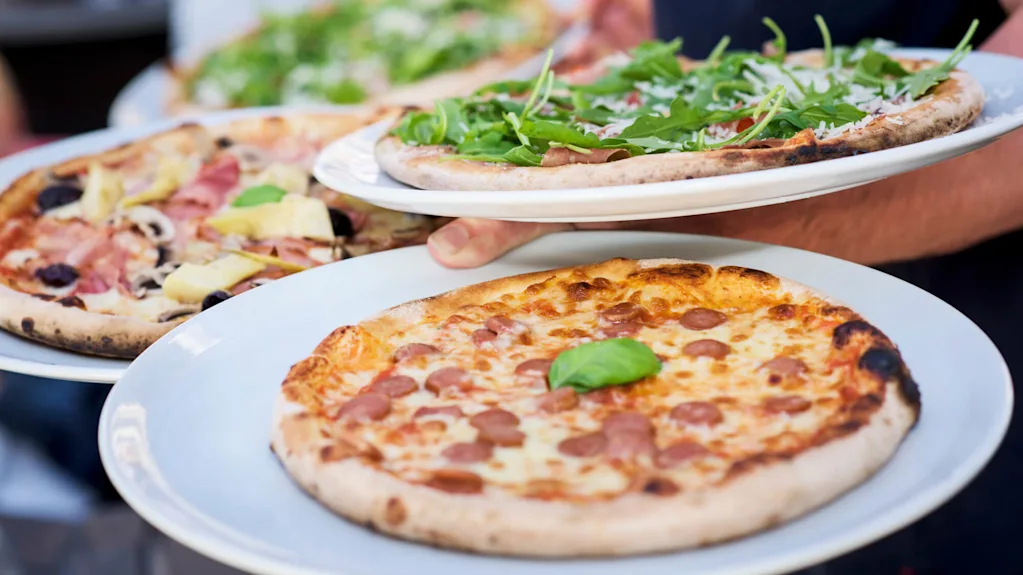 Neljä pizzaa valkoisilla lautasilla tarjoillaan, jokaisessa erilaisia täytteitä kuten basilikaa, rucolaa, sieniä ja leikkeleitä. Taustana ulkotilaa.