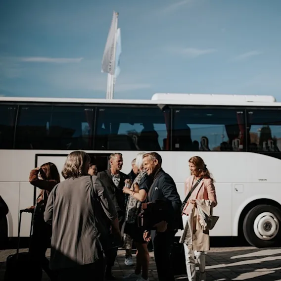 En grupp människor som pratar och ler utanför en vit buss under en klarblå himmel, möjligen före eller efter en resa.