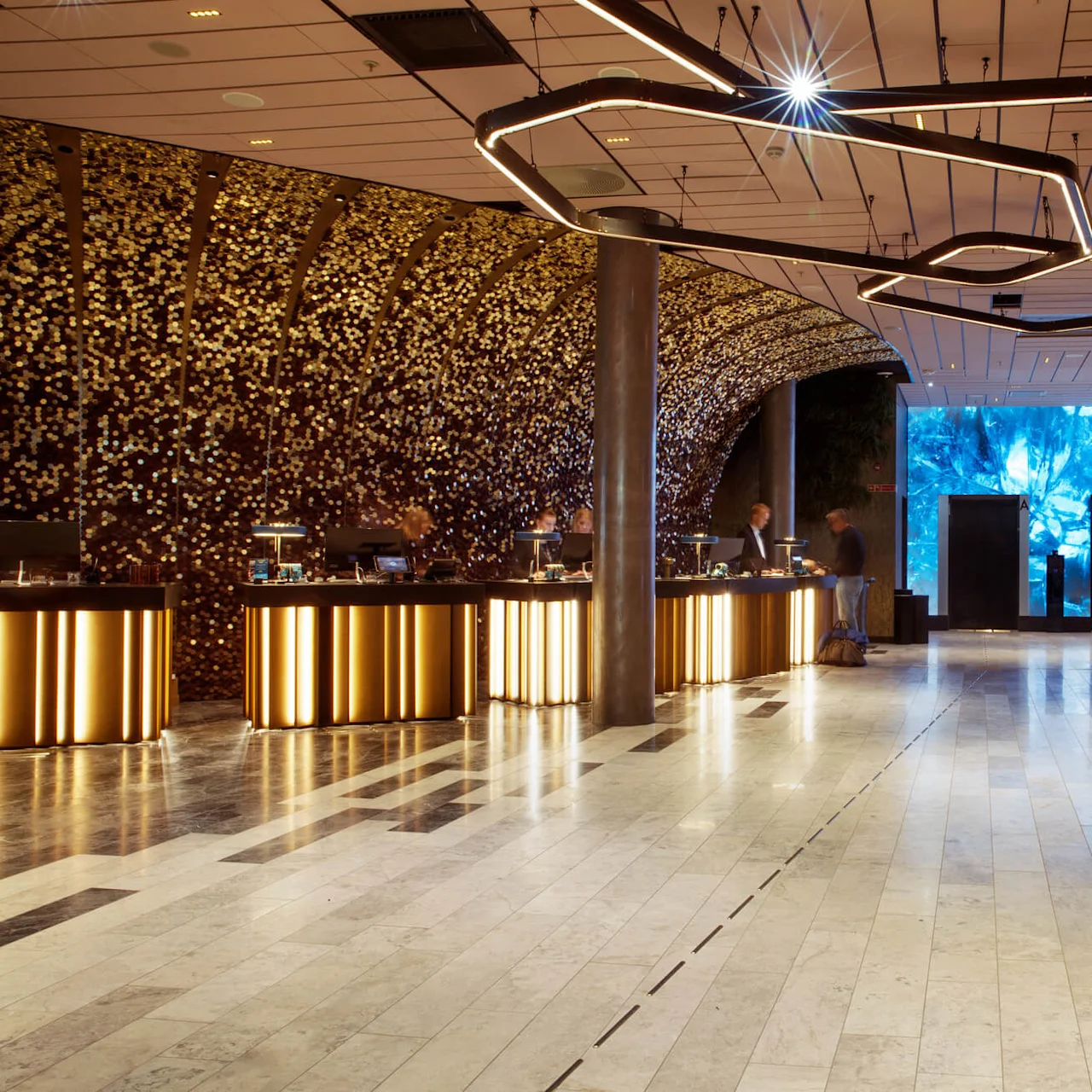 Reception area featuring illuminated counters where people interact, surrounded by a golden mosaic wall. Modern lighting hangs overhead, with elevators and blue-lit wall art in the background.