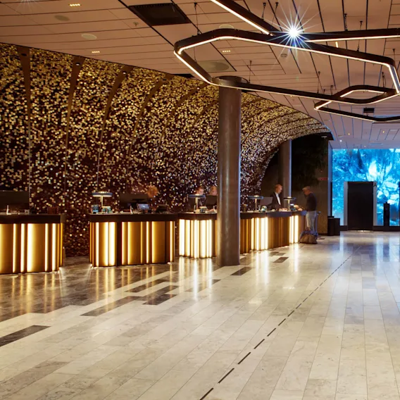 Reception area featuring illuminated counters where people interact, surrounded by a golden mosaic wall. Modern lighting hangs overhead, with elevators and blue-lit wall art in the background.