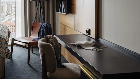 Et moderne hotellrom med skrivebord, stol og en lenestol ved et vindu, som antyder komfort og produktivitet.