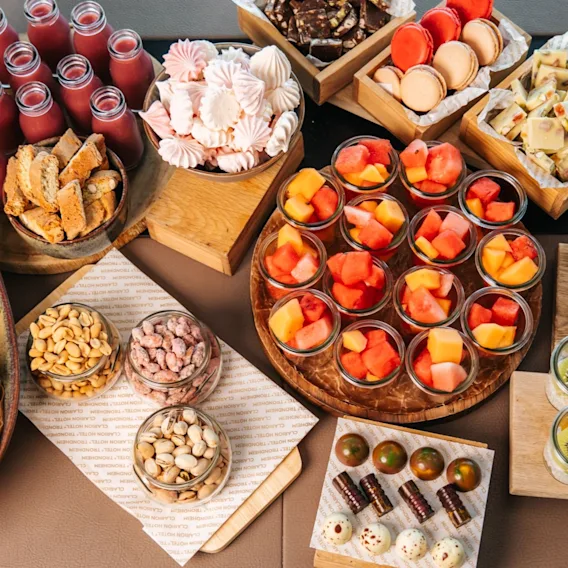 Et utvalg av matvarer vises på et bord, inkludert smørbrød, fruktkopper, makroner, nøtter og bakverk. Variasjonen presenteres i trebrett, krukker og glass, som skaper en fargerik buffé-setting.