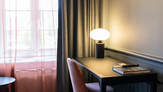 Et skrivebord med en lysende lampe står mod en væg, ved siden af et vindue med gennemsigtige gardiner. En stol er trukket ind, og en bog ligger på bordet.