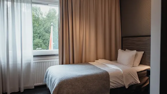 Viihtyisä hotellihuone, jossa on yhden hengen sänky, ikkuna vehreisiin puihin ja verhot rauhalliseen tunnelmaan.