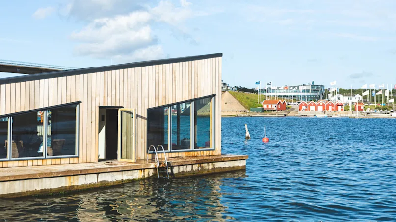 A wooden houseboat rests on calm water, with windows reflecting the blue sky, surrounded by a coastal town with red cabins, flags, and modern buildings.