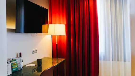 En väggmonterad tv ser ner på ett skrivbord med snacks och en flaska, bredvid ett rött draperi mjukt belyst av en stående lampa.