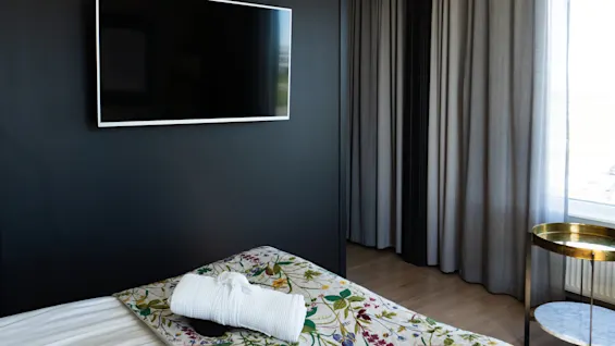 En TV är monterad på en mörk vägg bredvid en säng med ett blommigt överkast och hopvikt morgonrock. Grå gardiner täcker delvis ett fönster, och ett runt, gyllene sidobord står intill.