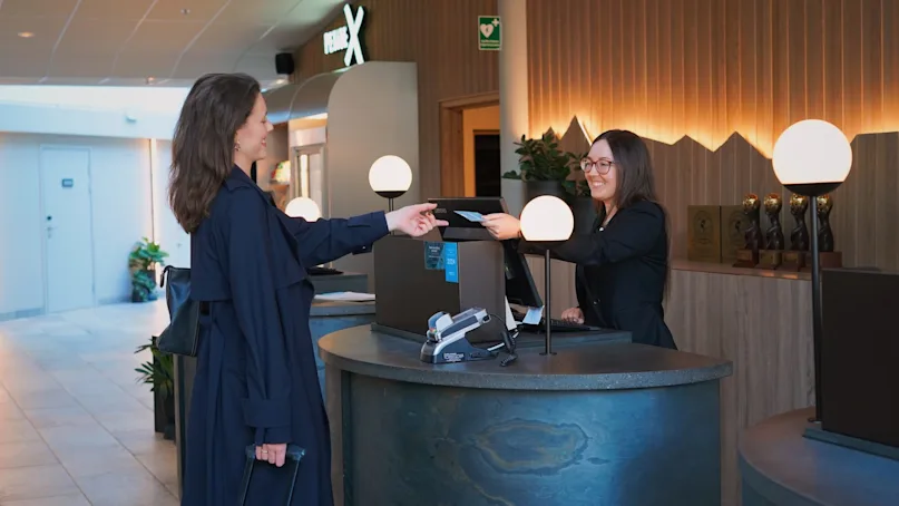 A woman in a blue coat hands a document to a smiling receptionist behind a stylish counter in a modern lobby, with decorative lighting and awards displayed.