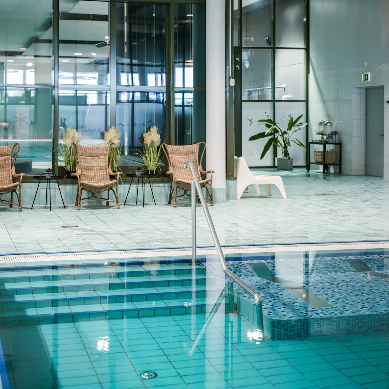 Swimming pool with turquoise tiles, surrounded by wicker chairs and plants. Large windows offer natural light. Signs read “Dampbadstue” near a door, and emergency exit. Relaxing indoor atmosphere.