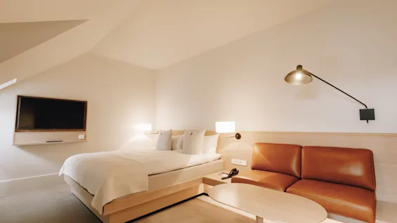 Ljust och modernt hotellrum med en stor säng, en brun skinnsoffa och ett litet soffbord under snedtak.