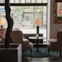 Fyra stoppade fåtöljer omger ett litet runt bord på en grön matta i ett mjukt upplyst café. Stora fönster visar utomhusskyltar: "APOTEK" och "GALLERIA DUVAN", med träd som syns utanför.