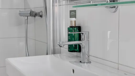 A white bathroom sink with a modern faucet, toiletries on a glass shelf above it. Clean, tiled environment.