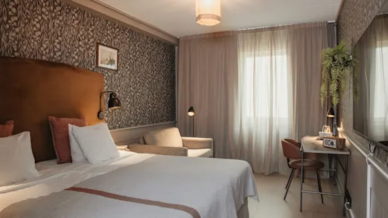 A cozy hotel room with a comfortable bed, a small seating area, and a desk by a window with sheer curtains.