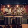 Cozy café setting, soft chairs facing round tables, surrounded by lush plants. Warm pendant lights hang from the ceiling, casting a gentle glow. Shelves with decorative items in the background.