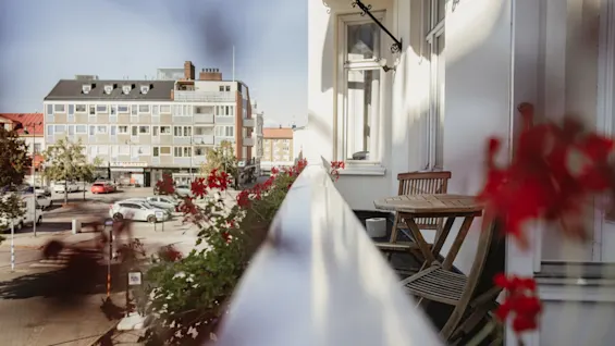 En hotellbalkong med röda blommor och ett litet träbord, med utsikt över en gata med byggnader och parkerade bilar.