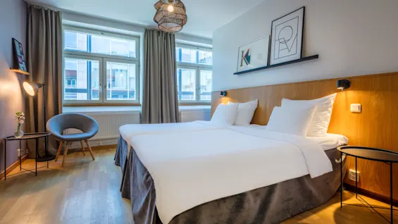 A neatly-made bed in a well-lit, modern hotel room with wooden headboard, artwork on the wall, and simplistic furniture.