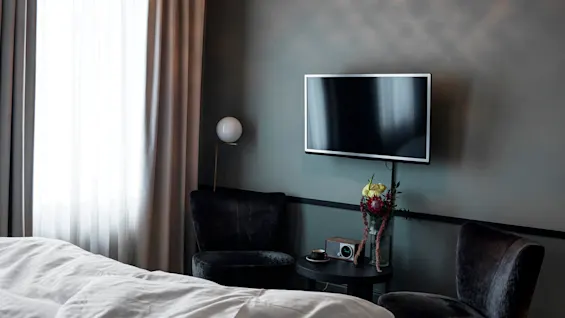 A wall-mounted TV in a modern bedroom displays a black screen, surrounded by velvet chairs, a circular table with a radio, flowers, and a softly lit window with curtains.