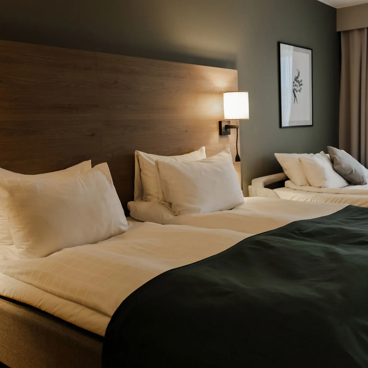 A double bed with white pillows and a dark blanket stands in a hotel room near a second bed. A lamp, picture frame, and curtains decorate the space.