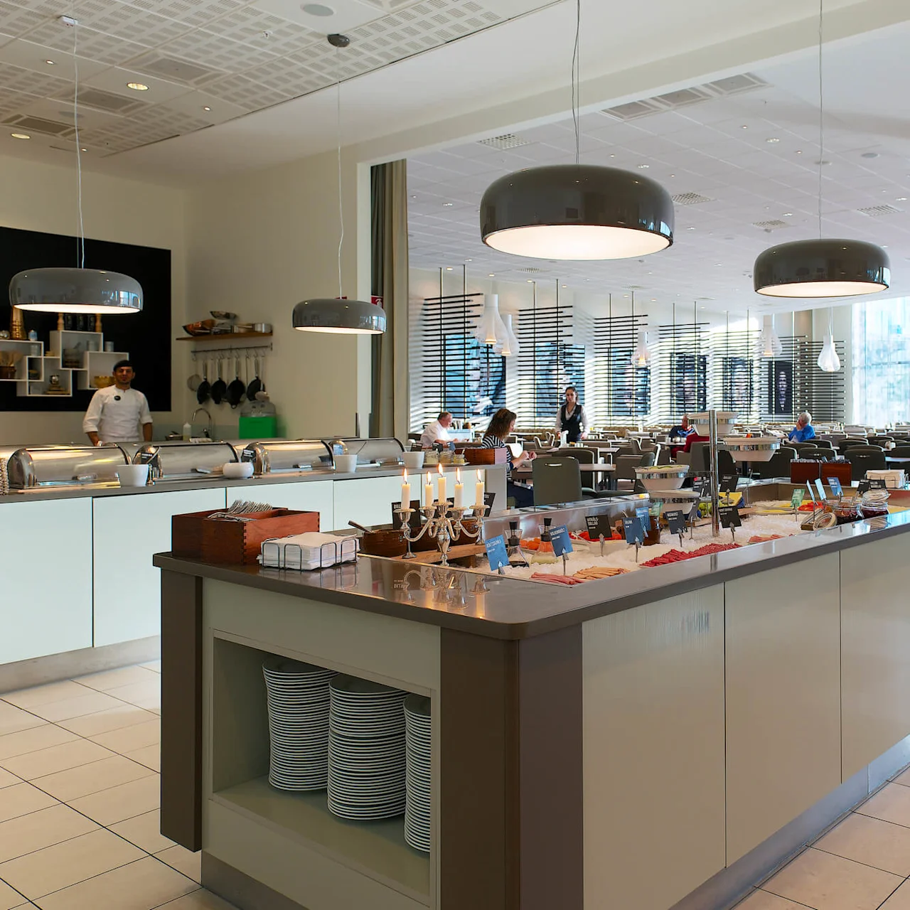 Buffet table displaying various foods in a spacious, modern restaurant dining area with overhead lights; people seated at tables in the background enjoy their meals.