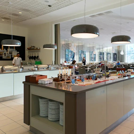 Buffébord med olika livsmedel i en rymlig, modern restaurangmatsal med taklampor; personer sitter vid bord i bakgrunden och njuter av sina måltider.