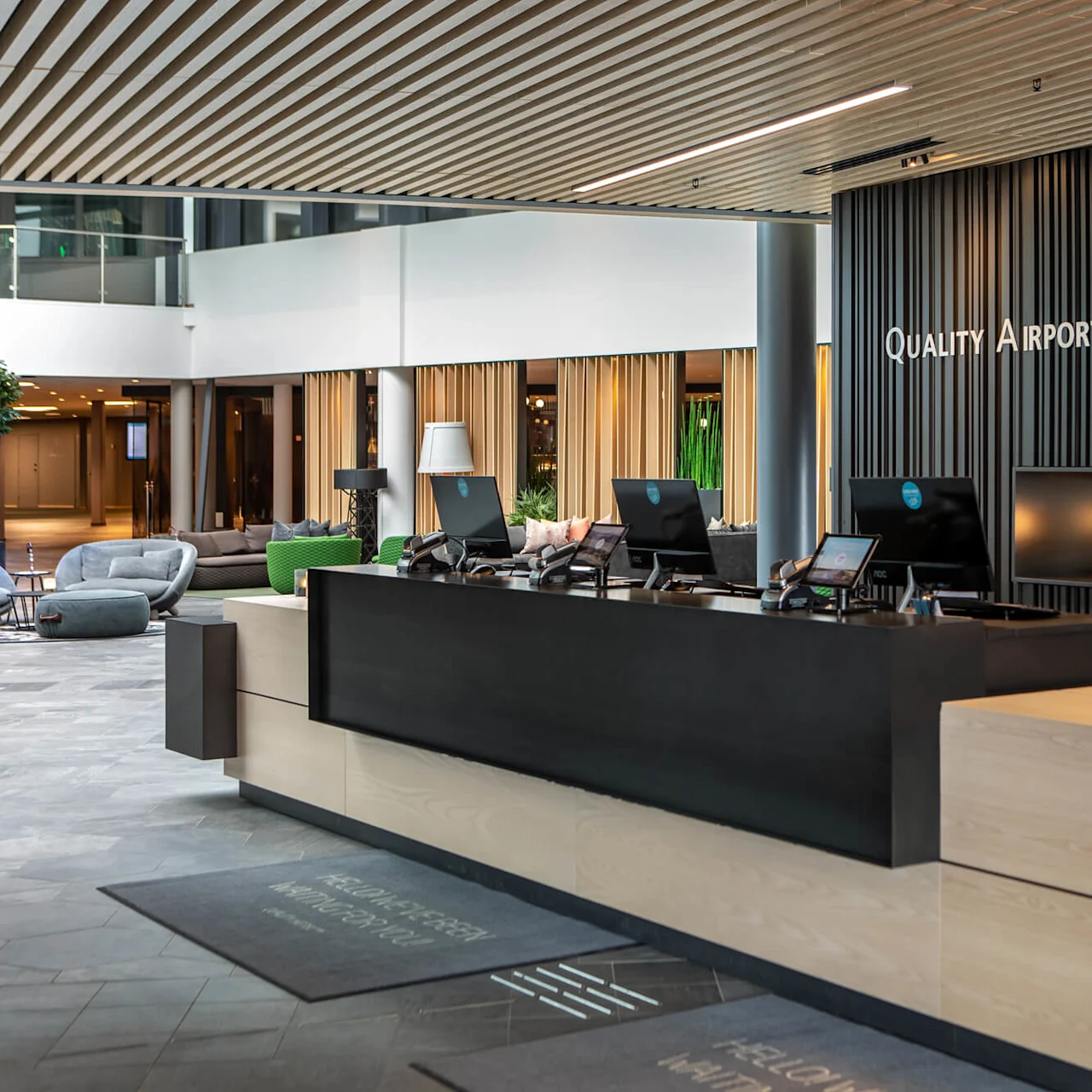 Reception desk with computers in a modern hotel lobby, featuring sleek design and branding. Text on the wall reads "Quality Airport Hotel Gardermoen."