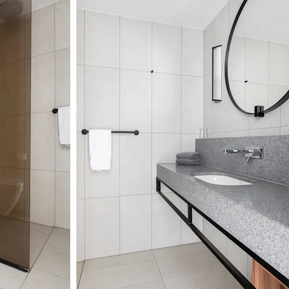 Wall-mounted toilet, shower, and sink station in a modern bathroom with light tiles and wooden accents. Minimalist design, featuring a round mirror and towel rack.