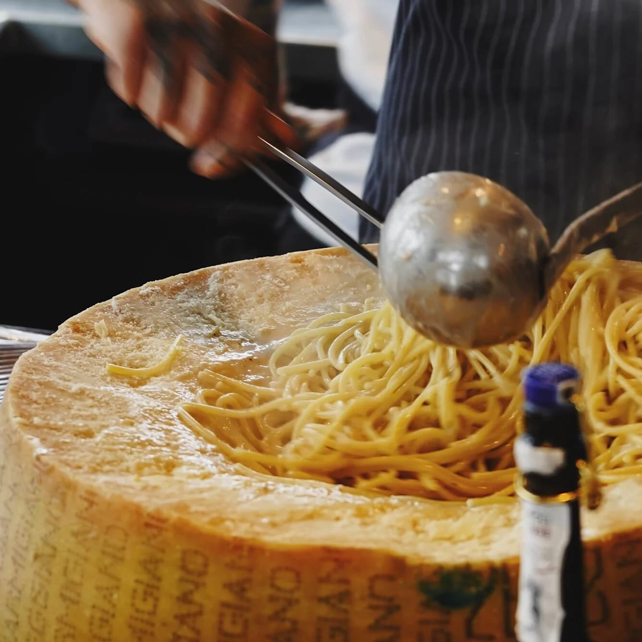 En osthjul holder dampende spaghetti som blandes med tang og øse; en persons hender er synlige, og "REGGIANO" er trykt på ostens overflate i et kjøkkenmiljø.