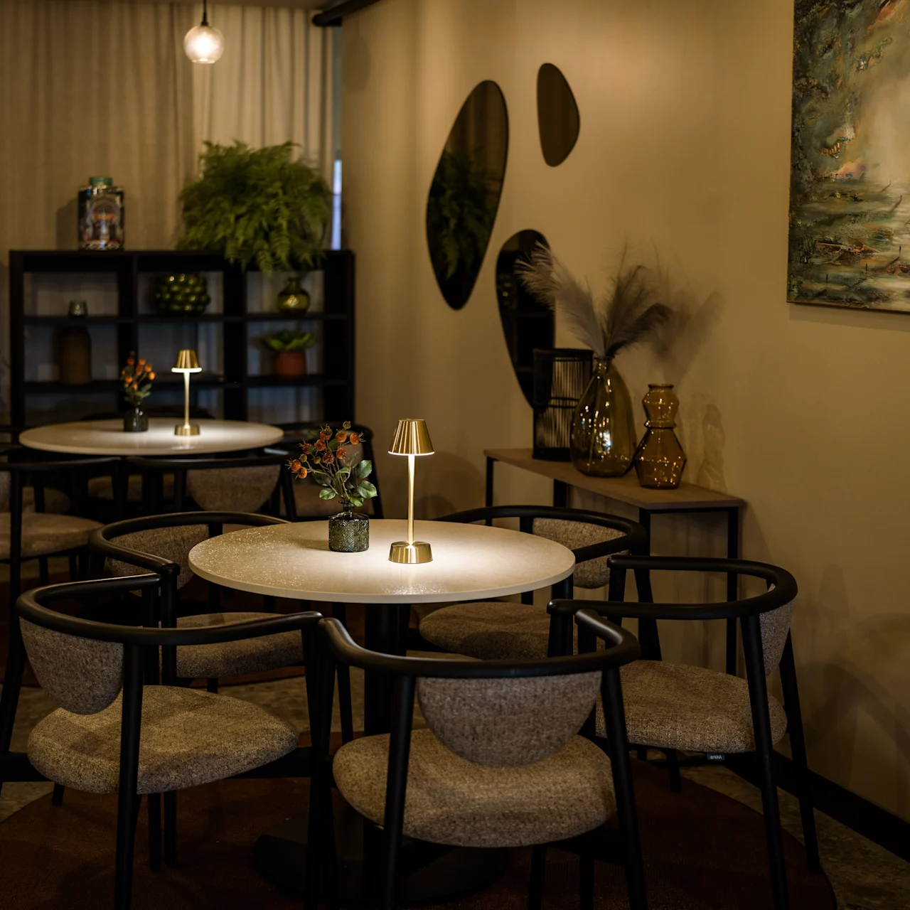 Tables with small lamps and vases sit in a warmly lit, cozy café. Surrounding shelves hold plants and decorative items, with artwork and wall mirrors enhancing the intimate ambiance.