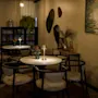 Tables with small lamps and vases sit in a warmly lit, cozy café. Surrounding shelves hold plants and decorative items, with artwork and wall mirrors enhancing the intimate ambiance.