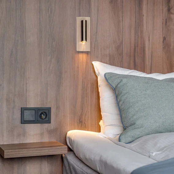 Et koselig hotell nattbordområde med en varm vegglampe, en komfortabel seng med puter og en praktisk stikkontakt.