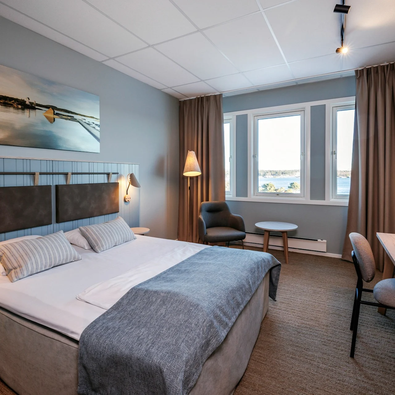 A neatly arranged hotel room features a large bed with grey accents, a desk, chair, and a window revealing a scenic view, adorned with modern lamps.