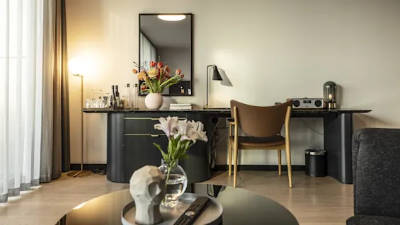 Et svart skrivebord med et speil holder en vase med blomster, bøker og en lampe; en stol er plassert i nærheten. Foran er det et salongbord med en hodeskalle-skulptur og flere blomster.